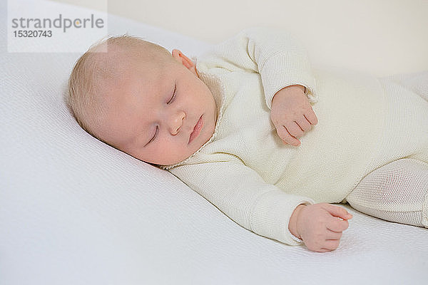 Junger Säugling in weißer Kleidung von 2 Monaten schläft auf einem weißen Bett.