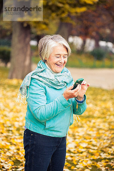 Blick auf eine lächelnde hübsche ältere Frau mit seinem Telefon auf gelben Blättern vor einem Baum mit herbstlichen Farben.