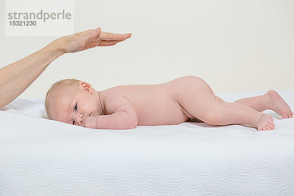 Nacktes  2 Monate altes Baby liegt auf dem Bauch auf einem weißen Bett und hat die Hand seiner Mutter über dem Kopf.