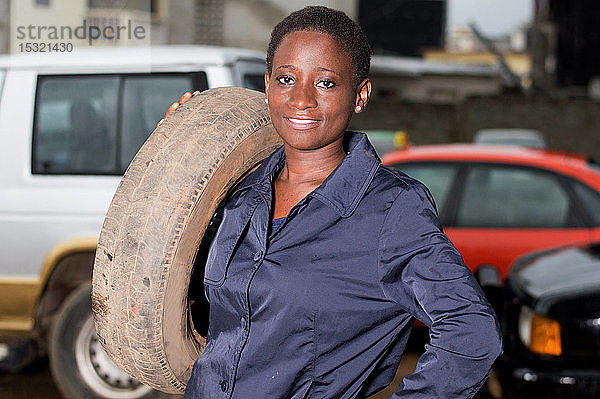 Junge Mechanikerin steht lächelnd mit einem Reifen auf der Schulter in ihrer Werkstatt.