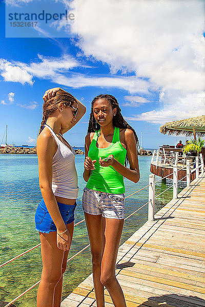 2 lächelnde Freundinnen sprechen auf einem Steg  der von einer Bar auf Stelzen an einem karibischen Strand in einer paradiesischen Landschaft kommt.