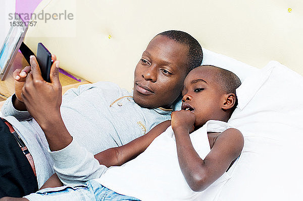 Ein Mann  der mit seinem Kind auf dem Bett liegt  zeigt ihm  wie man ein Mobiltelefon benutzt.