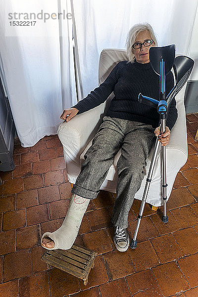 Frau auf einem Stuhl sitzend  mit einem eingegipsten Bein auf einer Fußstütze und mit Krücken in der Hand