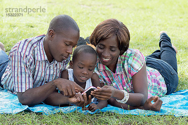 Diese Familie liegt auf einem Stück Stoff im Gras und konsultiert mit ihrem Kind ein Mobiltelefon.