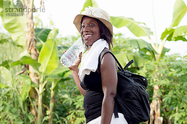 Diese lächelnde junge Frau  die eine Flasche Mineralwasser in der Hand hält und eine Tasche auf dem Rücken trägt  kehrt nach dem Training für den Radsport in die Stadt zurück.
