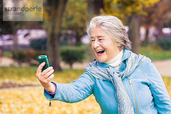 Blick auf eine lächelnde und ausdrucksstarke hübsche ältere Frau  die auf ihr Telefon auf gelben Blättern vor einem Baum mit herbstlichen Farben schaut.