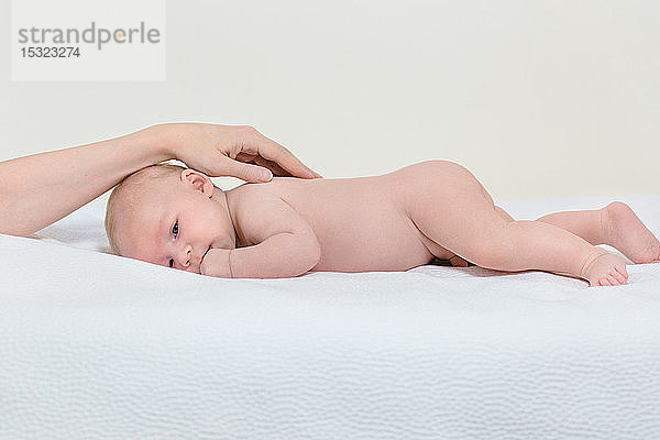 Hand einer Mutter auf dem Rücken ihres 2 Monate alten Babys  das auf dem Bauch in einem weißen Bett liegt.