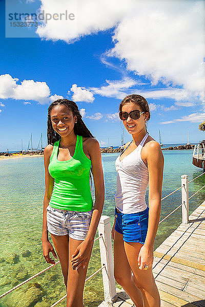 2 lächelnde Freundinnen posieren auf einem Steg  der von einer Bar auf Stelzen an einem karibischen Strand in einer paradiesischen Landschaft kommt.
