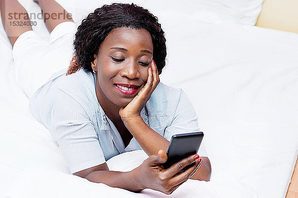 diese lächelnde junge Frau liegt auf einem Bett und sendet eine Nachricht auf einem Mobiltelefon