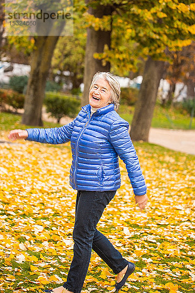 Porträt einer lächelnden und ausdrucksstarken älteren Frau  die mit einem dynamischen Schritt in einem Park vor gelben Blättern spazieren geht.