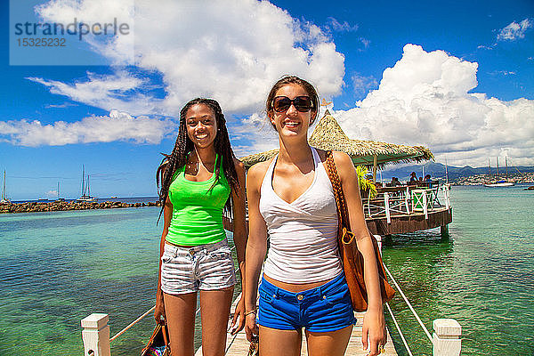 2 lächelnde Freundinnen  die auf einem Steg aus einer Bar auf Stelzen an einem karibischen Strand in einer paradiesischen Landschaft kommen.