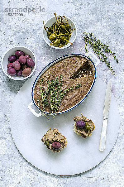 Leberpastete in Keramikform  dekoriert mit frischem Thymian und zwei Baguette-Sandwiches mit Pastete  Kapern  Oliven auf einem Marmorständer