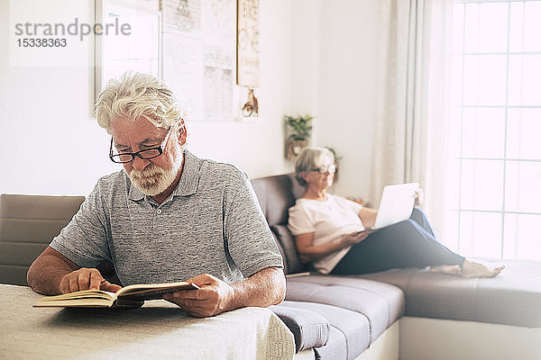 Älterer Mann liest ein Buch  während seine Frau im Wohnzimmer einen Laptop benutzt