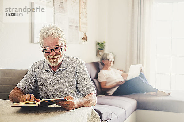Älterer Mann liest ein Buch  während seine Frau im Wohnzimmer einen Laptop benutzt