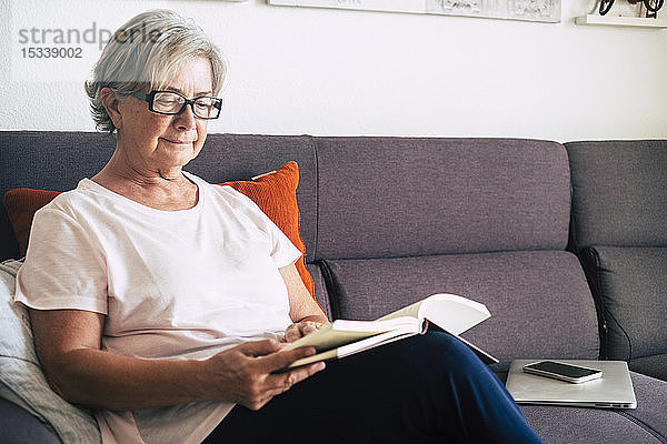 Ältere Frau liest ein Buch mit Laptop und Smartphone auf dem Sofa
