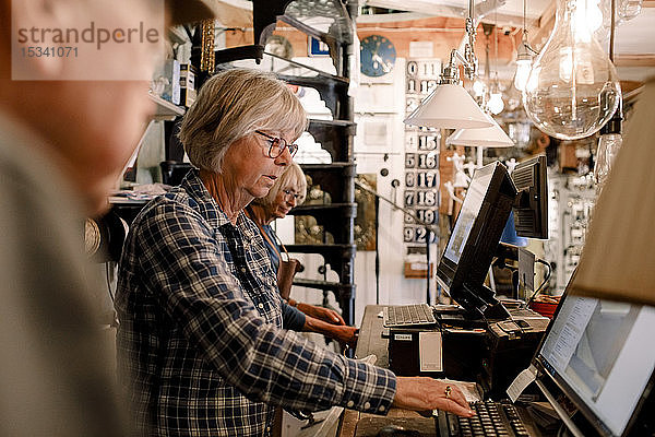 Selbstbewusste ältere Besitzerin  die einen Computer benutzt  während sie an der Kasse eines beleuchteten Eisenwarenladens inmitten von Kollegen steht
