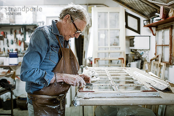 Seitenansicht eines älteren männlichen Eigentümers  der den Fensterrahmen auf der Werkbank in der Werkstatt kratzt