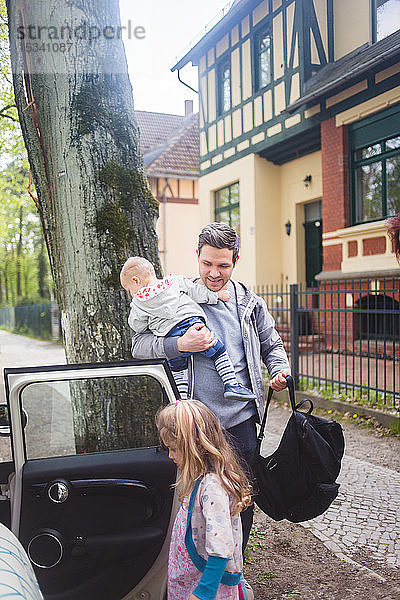 Vater trägt Sohn und Tasche  während er ein Mädchen beim Einsteigen ins Auto beobachtet