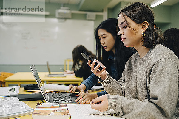 Gymnasiastin benutzt ein Smartphone  während sie im Klassenzimmer neben einer Klassenkameradin sitzt und am Laptop lernt