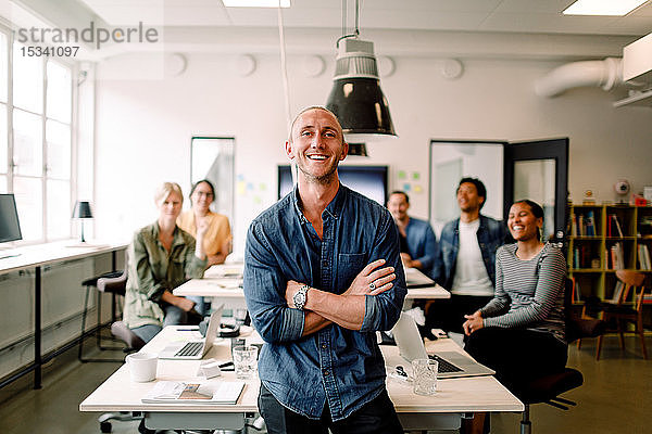 Porträt eines lächelnden männlichen Unternehmers stehend mit verschränkten Armen und Angestellten am Schreibtisch sitzend
