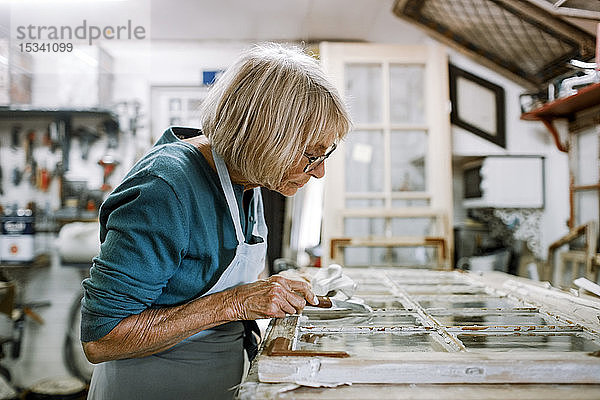 Seitenansicht eines älteren weiblichen Eigentümers  der Fensterrahmen auf der Werkbank in einer Werkstatt schabt