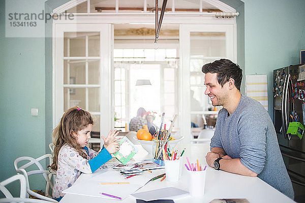 Lächelnder Vater schaut ein Mädchen an  das ein Bilderbuch in der Küche hält