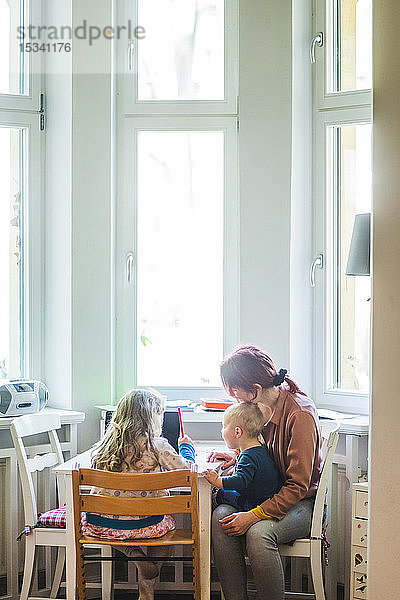 Mutter sitzt mit ihrem Sohn zusammen  während sie einem Mädchen beim Lernen am Tisch assistiert