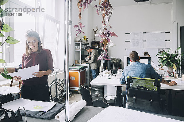Kreative Geschäftsfrau prüft Papiere  während Kollegen im Hintergrund im Büro arbeiten