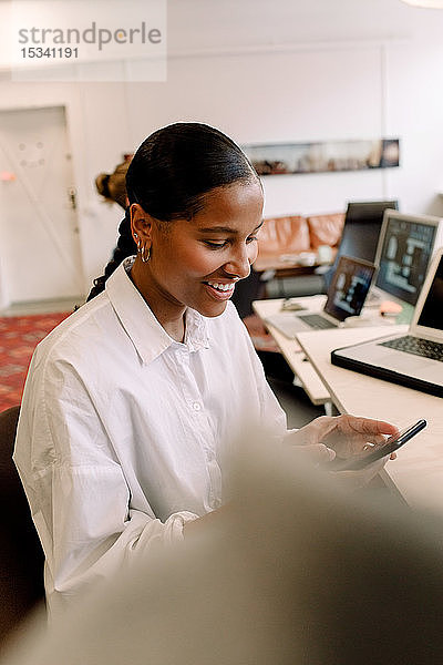 Lächelnde Geschäftsfrau benutzt Mobiltelefon am Schreibtisch im Büro