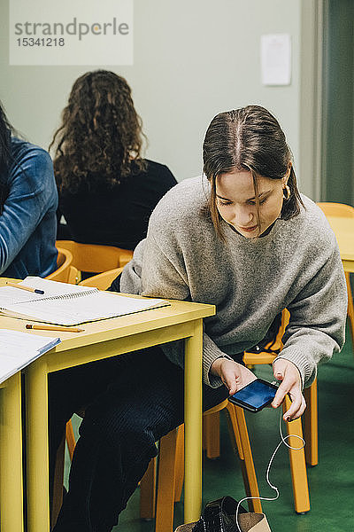 Teenager-Mädchen benutzt Smartphone  während sie im Klassenzimmer am Schreibtisch sitzt