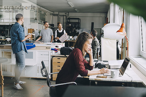 Kreative Geschäftsfrau mit Laptop am Schreibtisch  während Kollegen im Hintergrund arbeiten