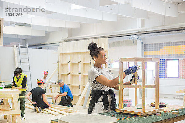Zuversichtliche junge Tischlerin reibt in beleuchteter Werkstatt Sandpapier auf Holztisch an Werkbank