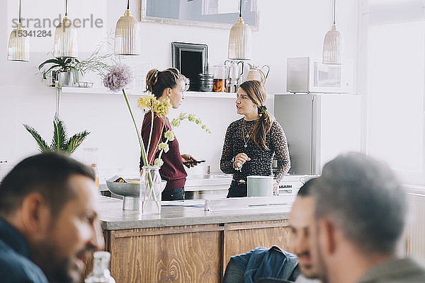 Kreative Unternehmerinnen diskutieren auf der Kücheninsel  während Kollegen im Büro im Vordergrund stehen