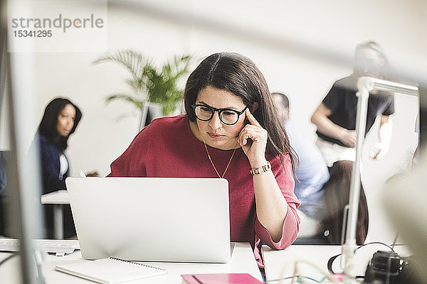 Selbstbewusste Geschäftsfrau benutzt Laptop am Schreibtisch  während sie an einem kreativen Arbeitsplatz sitzt