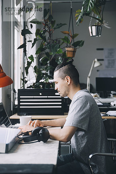 Seitenansicht eines selbstbewussten männlichen Hipsters mit Laptop am Schreibtisch im Kreativbüro