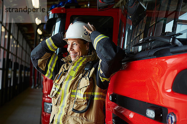 Lächelnde weibliche Feuerwehrfrau mit Helm  während sie in der Feuerwache beim Löschfahrzeug steht