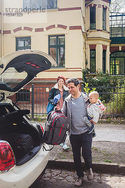 Frau sieht Mann an  der einen Sohn trägt  während sie Gepäck in den Kofferraum eines Autos legt