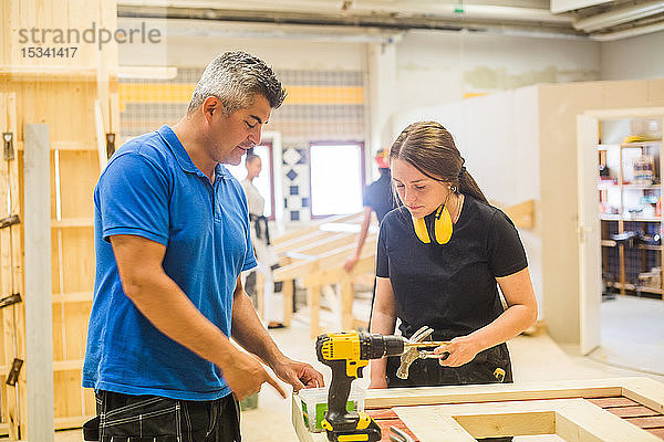 Männlicher Ausbilder unterrichtet weibliche Auszubildende an der Werkbank in beleuchteter Werkstatt