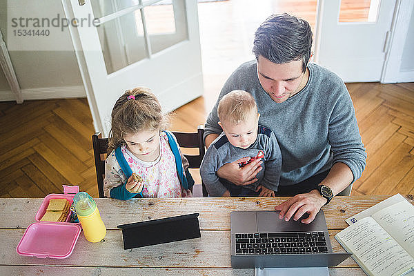 Hochwinkelansicht eines Vaters  der am Laptop arbeitet  während er mit dem süßen Sohn von der Tochter zusammensitzt und ein digitales Tablett am Tisch benutzt