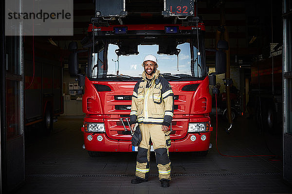 Ganzfigur eines lächelnden männlichen Feuerwehrmannes  der in der Feuerwache vor dem Löschfahrzeug steht