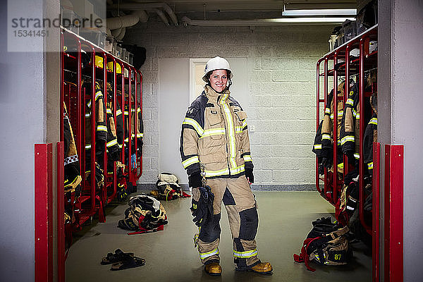 Porträt einer lächelnden Feuerwehrfrau in voller Länge  die in der Umkleidekabine der Feuerwache steht