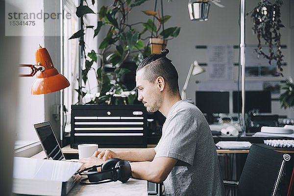 Seitenansicht eines männlichen Hipsters mit Laptop am Schreibtisch im Kreativbüro