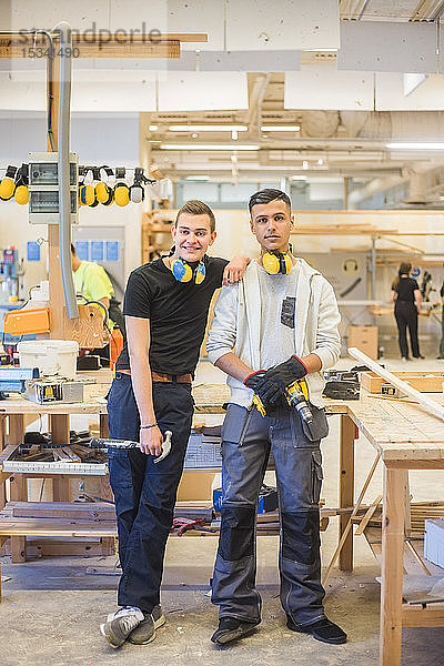 Lächelnder junger männlicher Auszubildender in voller Länge  der sich auf die Schulter eines Mitarbeiters stützt  während er in der Werkstatt Arbeitswerkzeuge hält