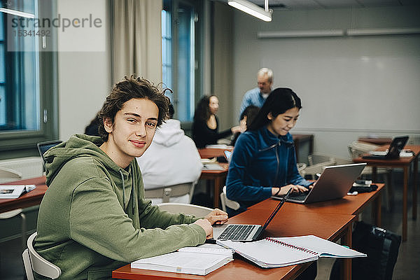 Porträt eines lächelnden Teenagers mit Laptop am Schreibtisch mit Klassenkameraden  die im Klassenzimmer lernen