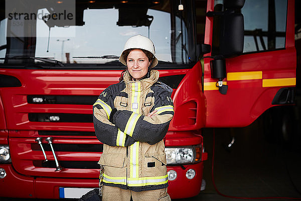 Porträt einer selbstbewussten Feuerwehrfrau  die mit verschränkten Armen gegen ein Feuerwehrauto auf der Feuerwache steht