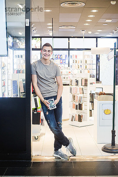 Porträt eines lächelnden jungen Verkäufers in voller Länge  der am Eingang des Ladens steht und die Telefonhülle hält