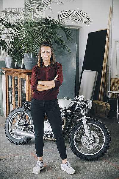 Porträt einer selbstbewussten Unternehmerin  die mit verschränkten Armen gegen ein Motorrad im Kreativbüro steht