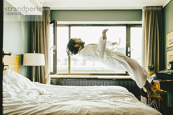 Verspielter Mann in Robe  der im Hotel auf dem Bett hüpft
