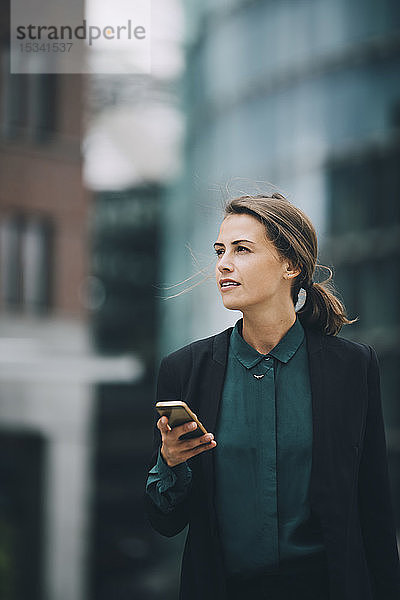 Selbstbewusste Geschäftsfrau schaut weg  während sie in der Stadt ein Smartphone in der Hand hält