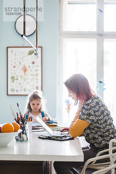 Seitenansicht einer mittleren erwachsenen Frau  die am Laptop arbeitet  während sie mit einem Mädchen auf der Kücheninsel sitzt